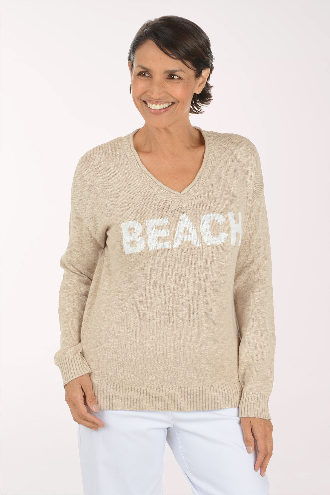 A woman wearing a beige V-neck knit sweater with the word 'BEACH' in large white letters across the chest, paired with white pants, against a plain light background.