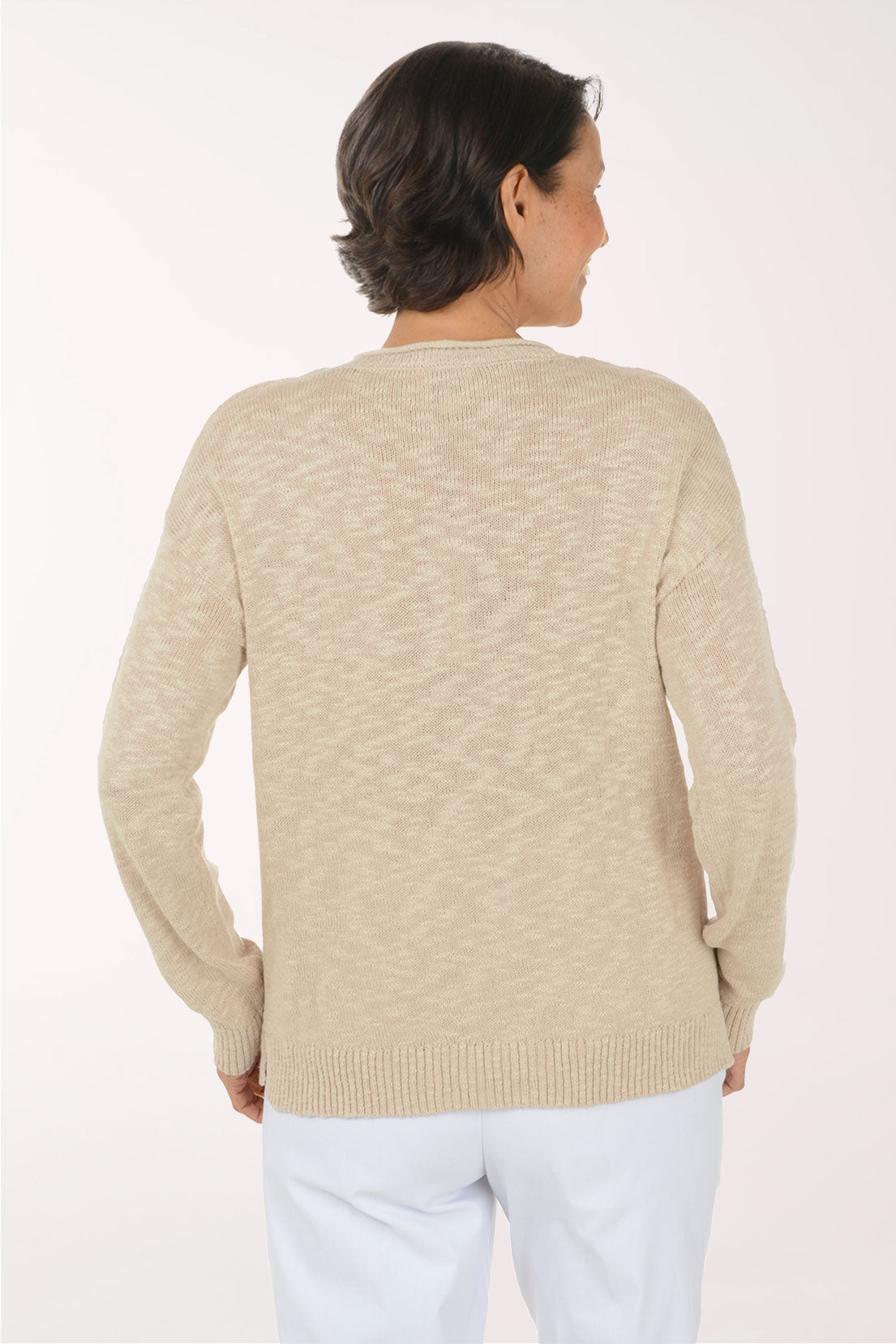 A woman wearing a beige V-neck knit sweater with the word 'BEACH' in large white letters across the chest, paired with white pants, against a plain light background.