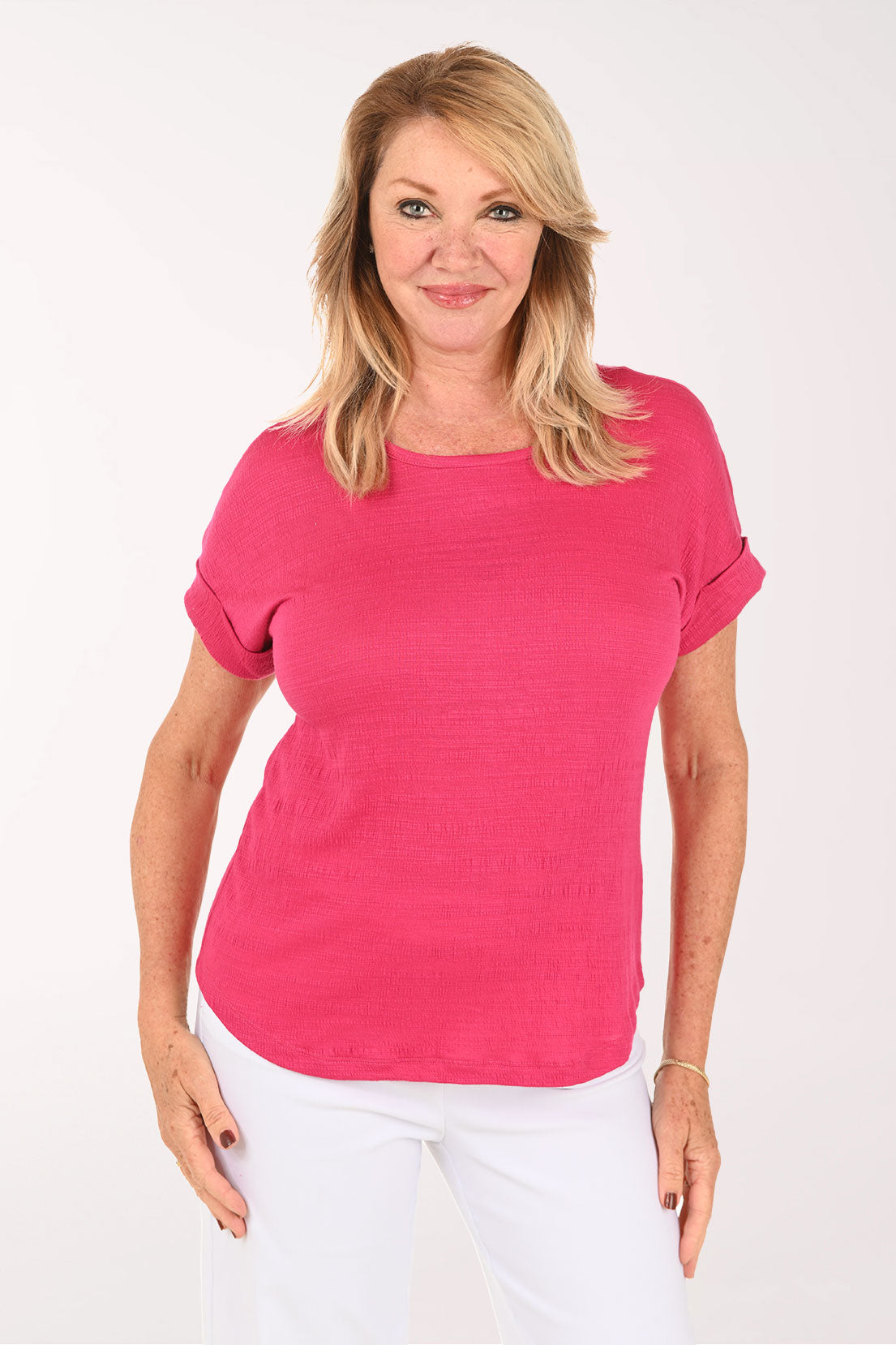 A smiling blonde woman wearing a bright pink short-sleeve crewneck top and white pants, standing against a plain white studio background.