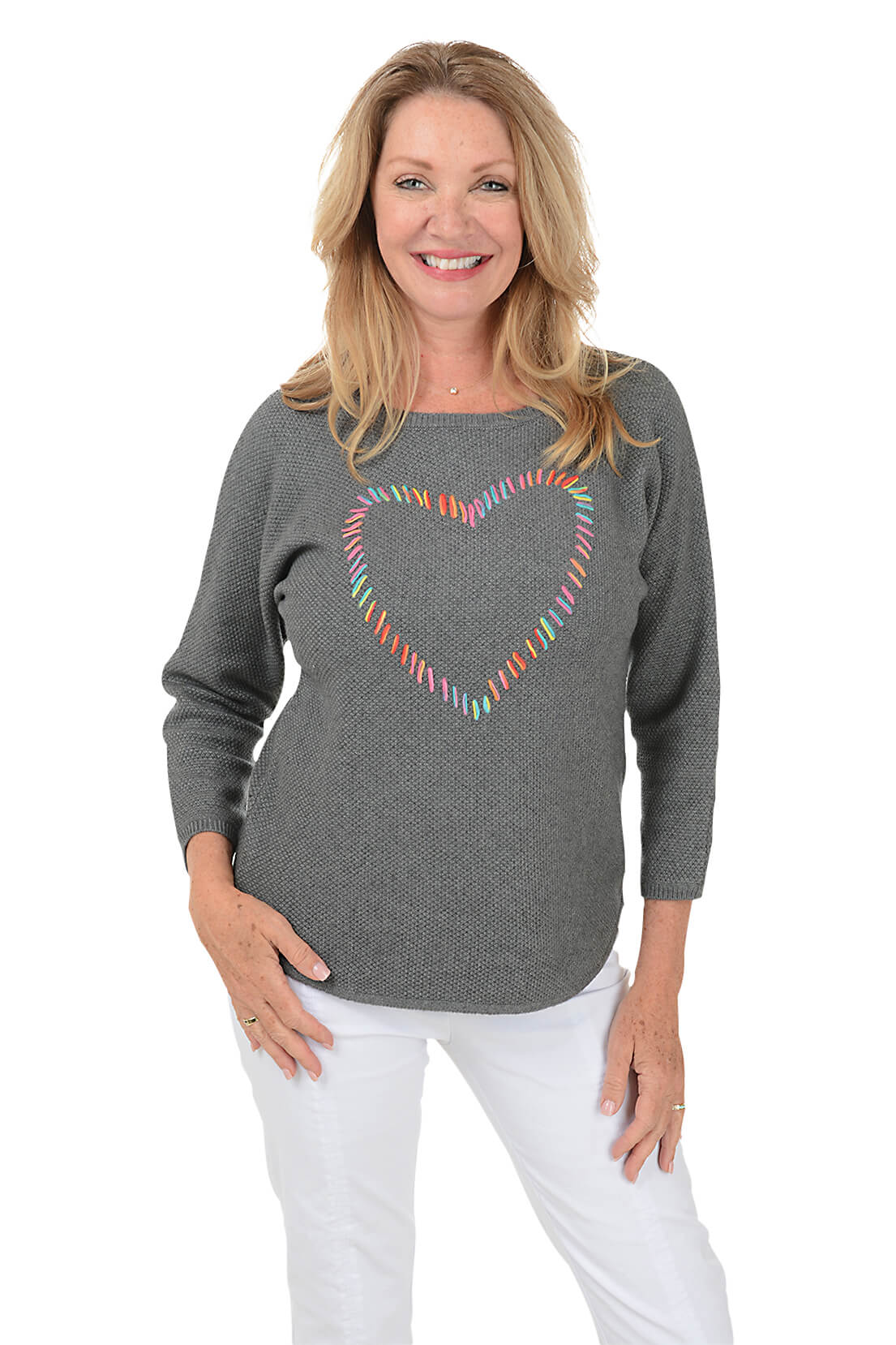 A smiling blonde woman wearing a heather gray long-sleeve top with a colorful heart outline graphic on the chest, paired with white pants, against a plain white background.
