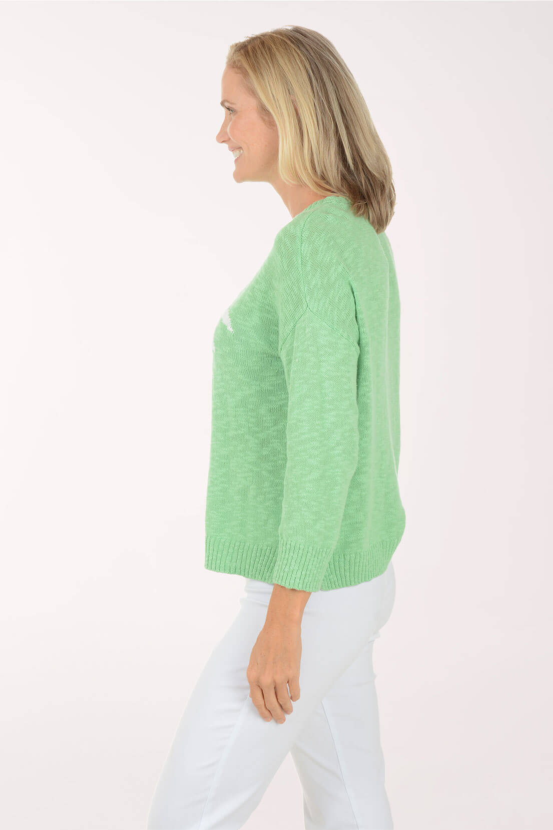 A woman shown in side profile wearing a light green knit sweater and white pants against a plain light background.