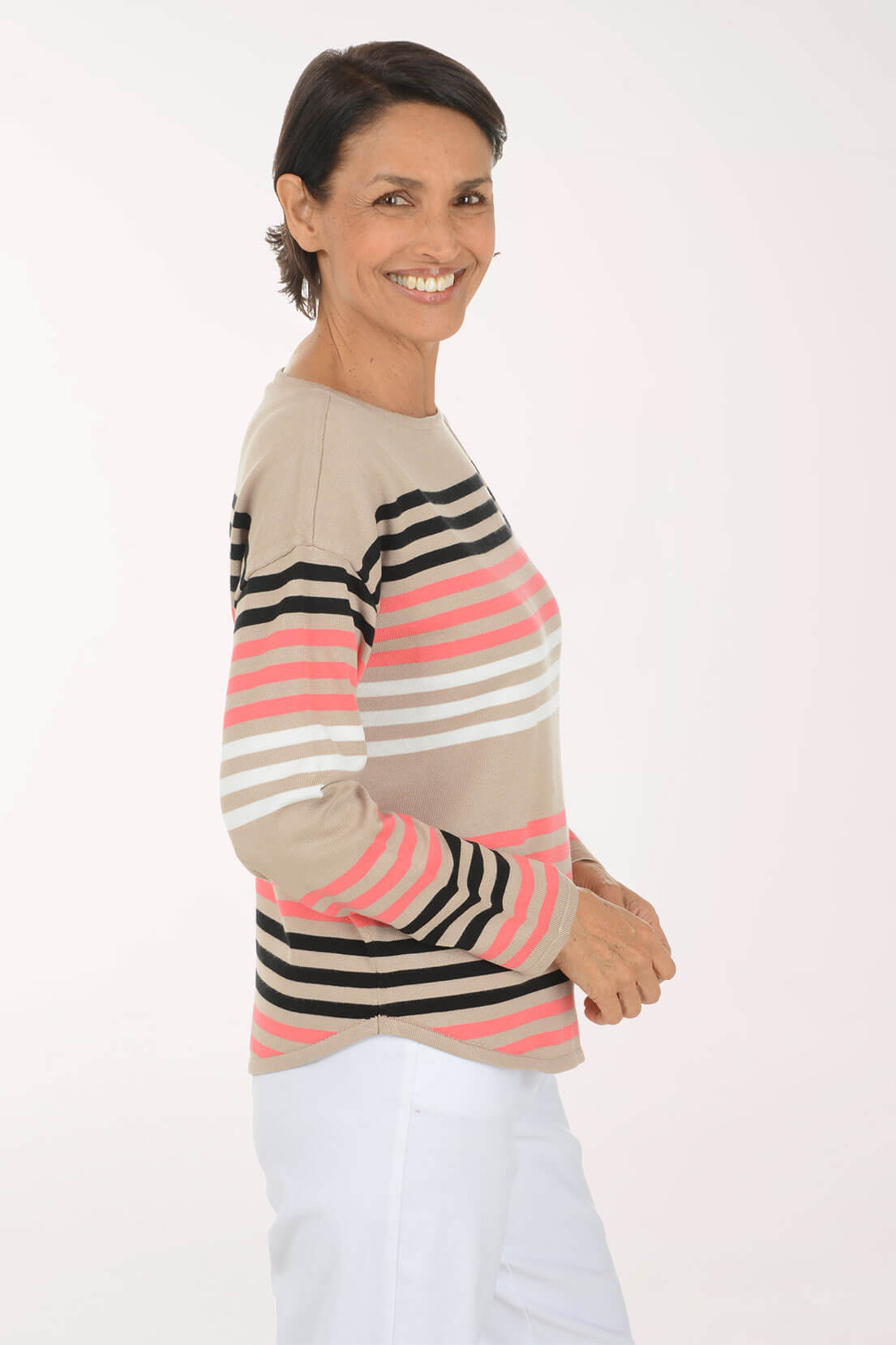A woman wearing a beige long-sleeve knit sweater with horizontal stripes in black, white, and pink, paired with white pants, photographed against a plain white studio background.