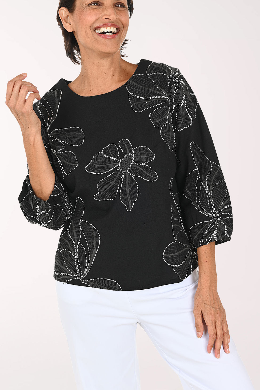 A smiling woman wearing a black three-quarter sleeve top with white floral embroidery, paired with white pants, against a plain white background.