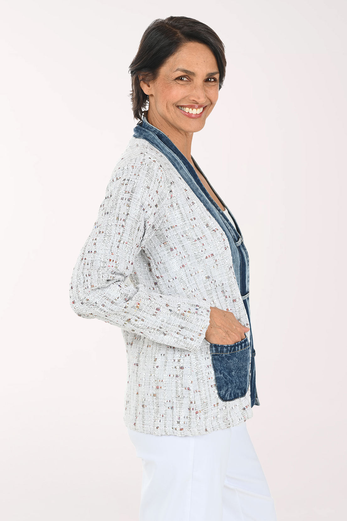 A smiling woman wearing a light gray speckled open-front cardigan with dark blue denim-style trim and front pockets, paired with a white top and white pants, against a plain light background.
