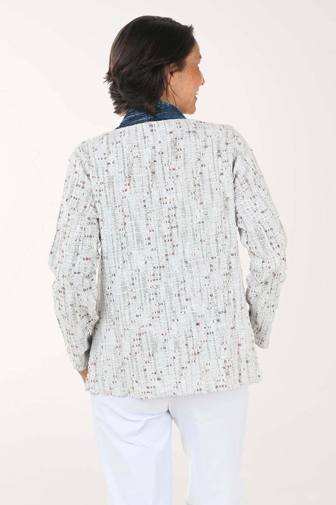 A smiling woman wearing a light gray speckled open-front cardigan with dark blue denim-style trim and front pockets, paired with a white top and white pants, against a plain light background.