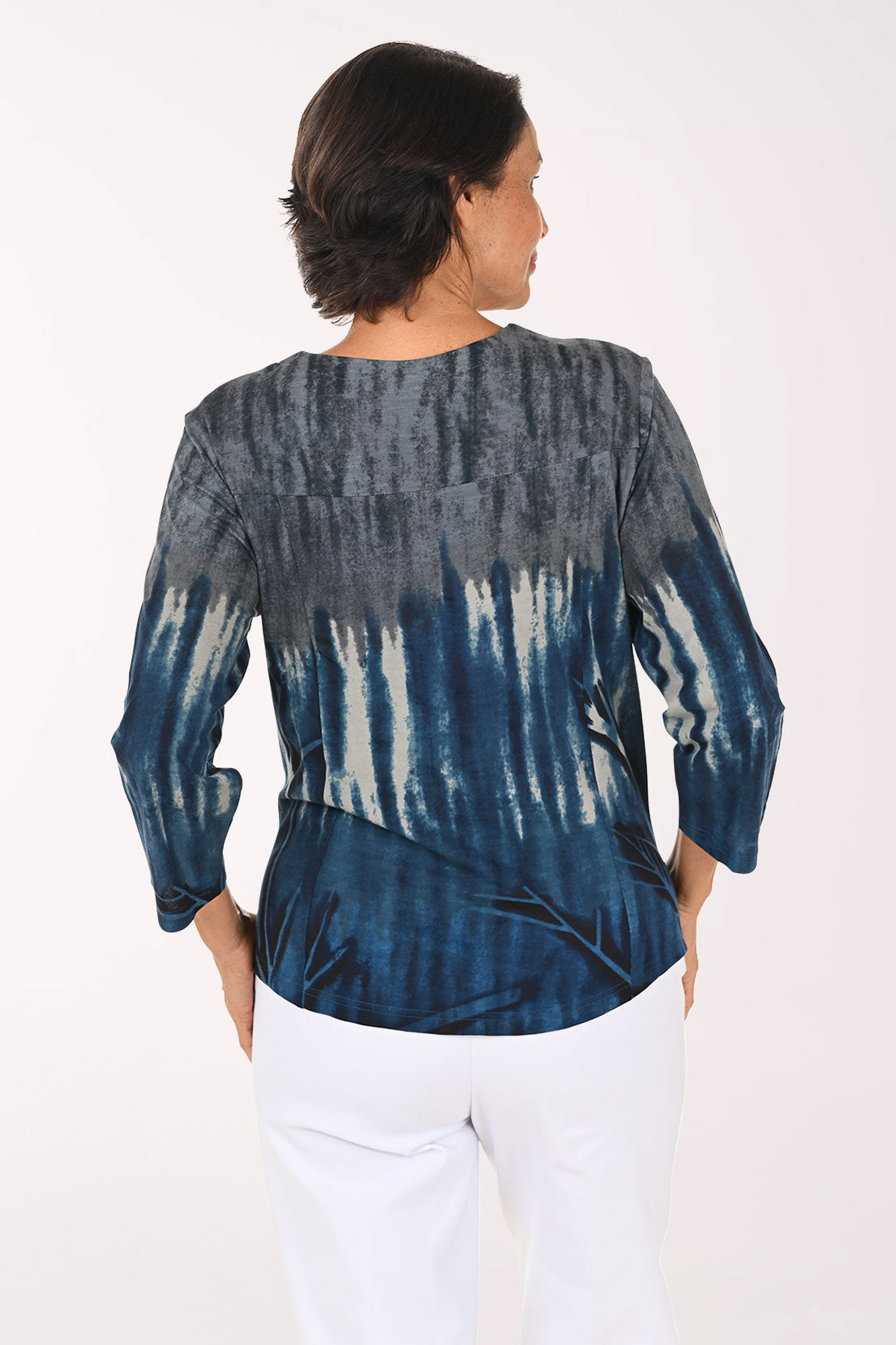 A smiling woman wearing a blue and gray tie-dye long-sleeve top with white pants, standing against a plain white studio background.