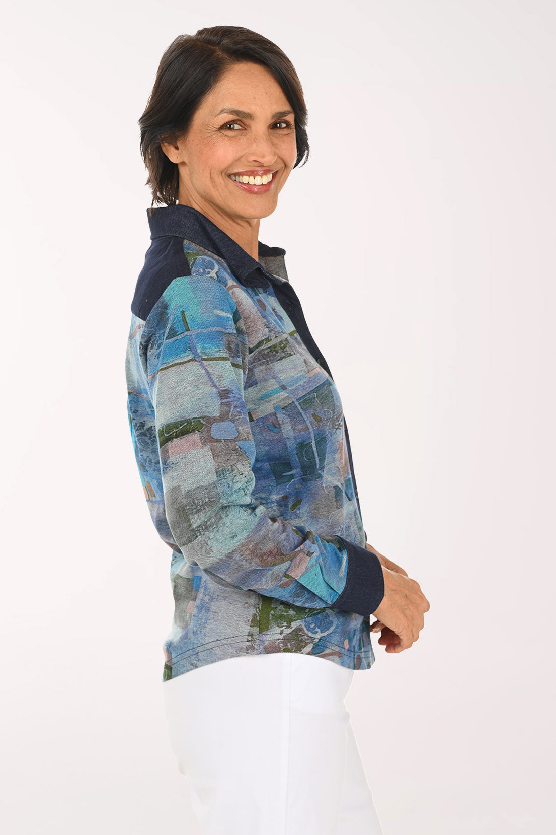 A smiling woman wearing a long-sleeve button-up shirt with an abstract blue, gray, and green patchwork print, paired with white pants, against a plain light background.