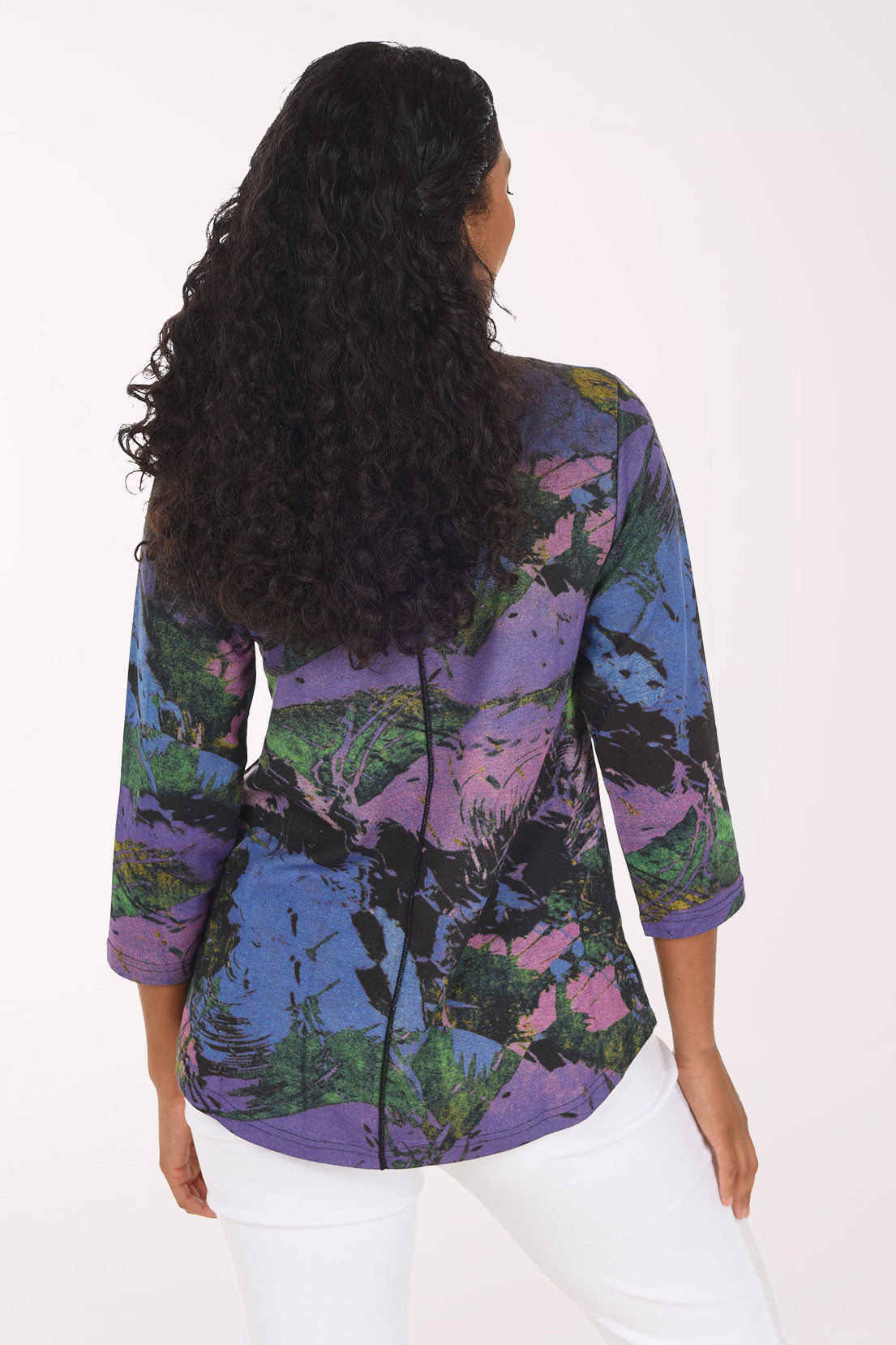 A smiling woman with long dark curly hair wearing a colorful abstract print top with three-quarter sleeves and white pants, standing against a plain white background.