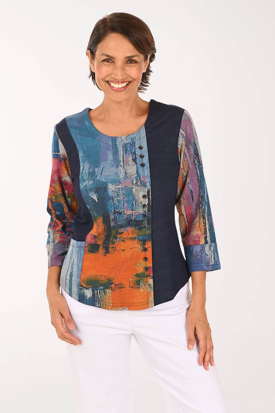 A woman modeling a three-quarter sleeve top with a multicolor abstract print on the front and sleeves, paired with white pants, against a plain white background.