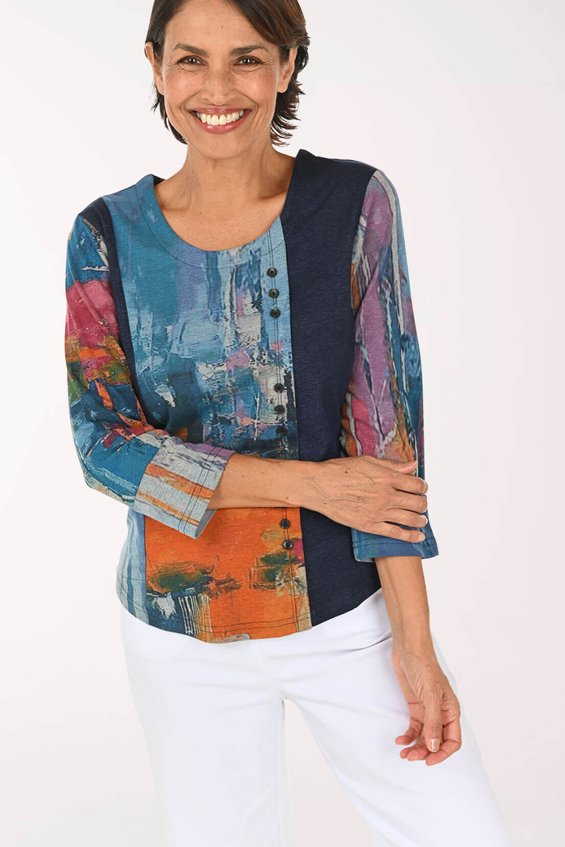 A woman wearing a three-quarter sleeve top with a multicolor abstract print and navy blue side panels, paired with white pants, against a plain white background.