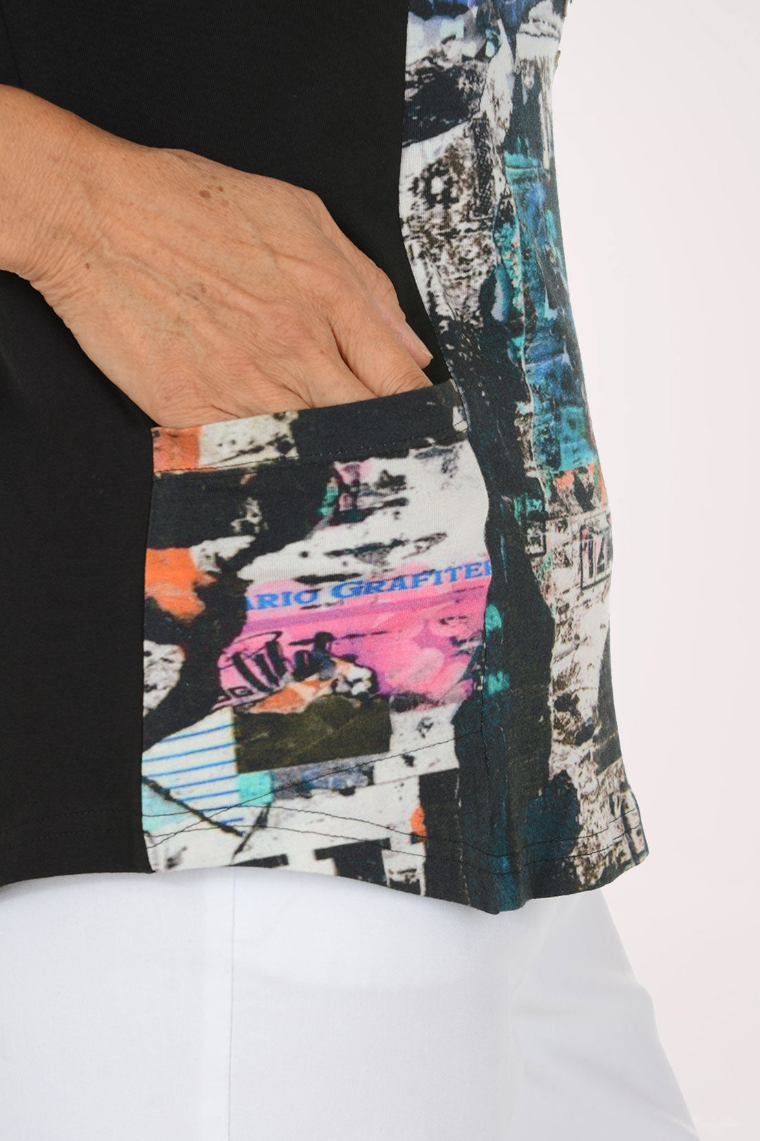 A woman modeling a three-quarter sleeve top with a black side panel and a multicolor abstract collage print on the front and sleeves, paired with white pants against a plain white background.