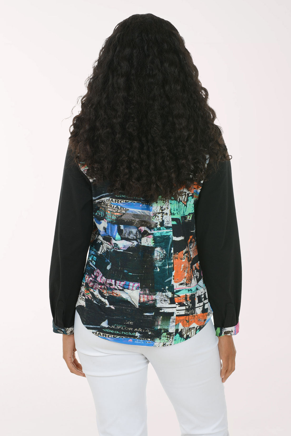 A woman modeling a long-sleeve button-up shirt with black sleeves and a colorful abstract collage print on the front panel, paired with white pants against a plain white background.