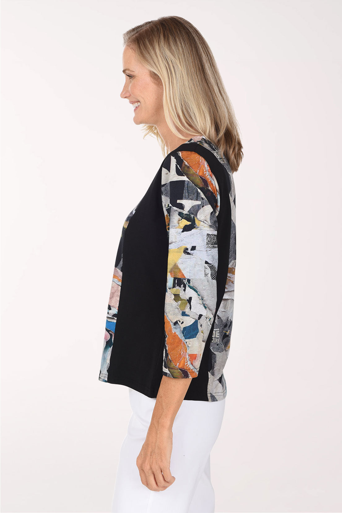 A woman wearing a long-sleeve collared blouse with a black side panel and a multicolor abstract collage print on the rest of the body, paired with white pants against a plain light background.