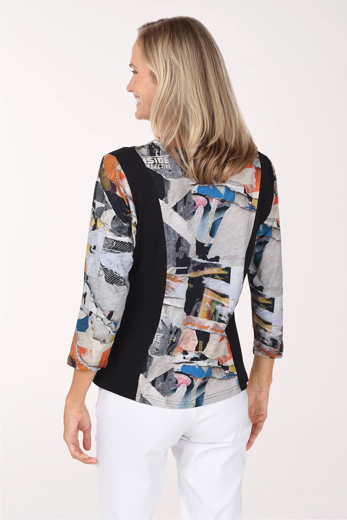 A woman wearing a long-sleeve collared blouse with a black side panel and a multicolor abstract collage print on the rest of the body, paired with white pants against a plain light background.