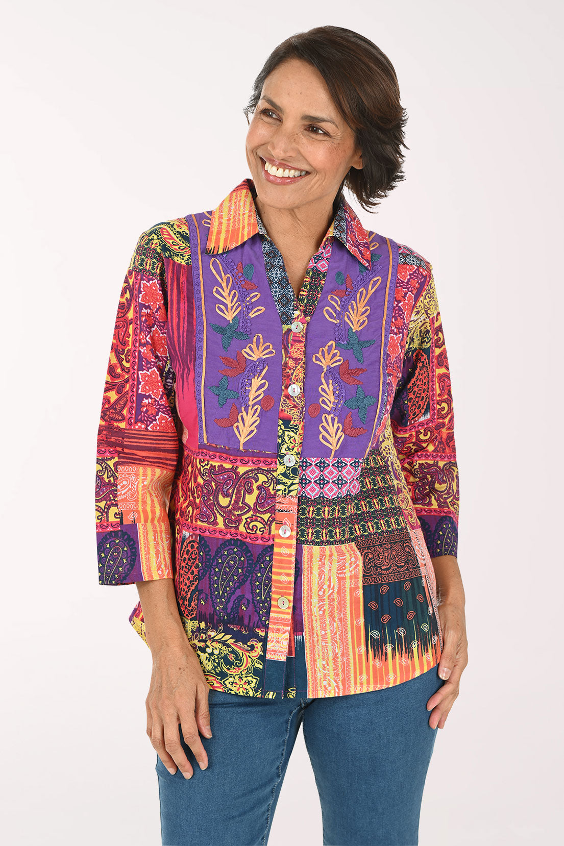 A woman modeling a vibrant, multicolored patchwork button-up shirt with intricate botanical and geometric patterns, paired with blue jeans against a plain white background.