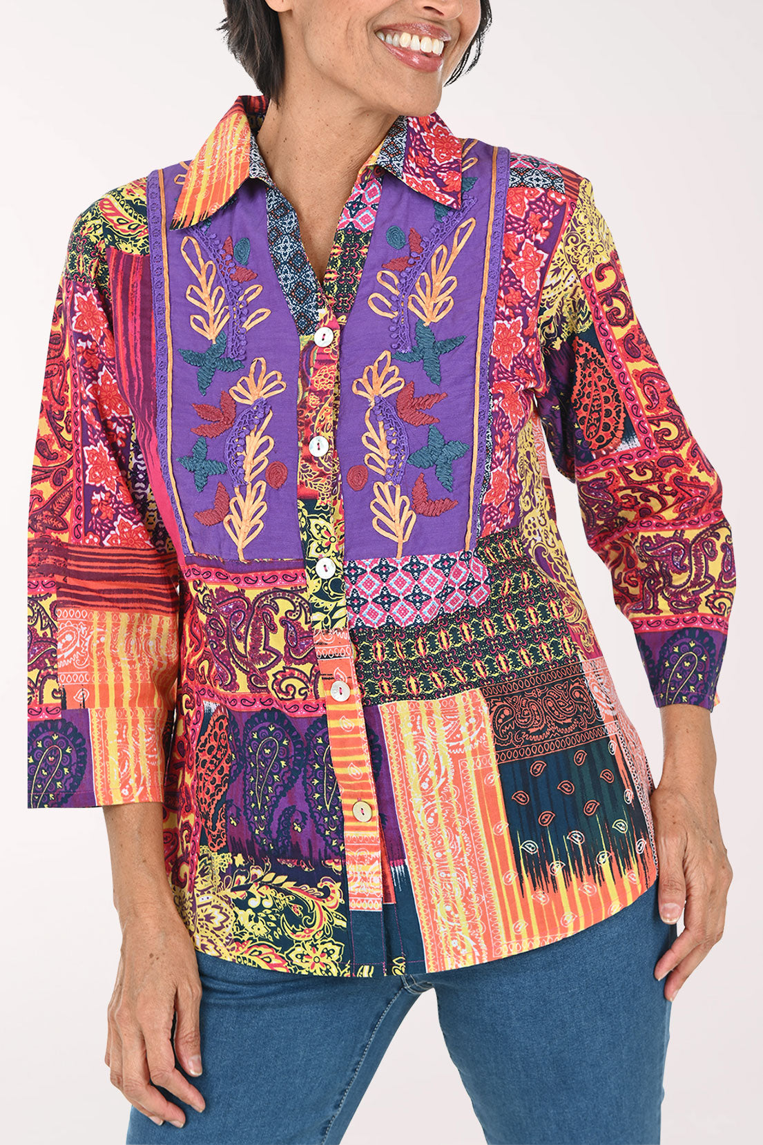 A woman modeling a vibrant, multicolored patchwork button-up shirt with intricate botanical and geometric patterns, paired with blue jeans against a plain white background.