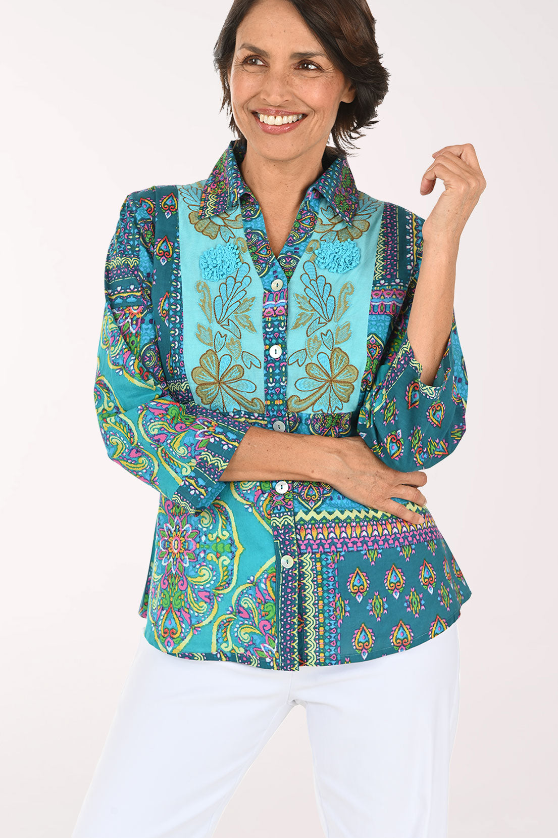 A smiling woman wearing a vibrant, multicolored patchwork print blouse with three-quarter sleeves and white pants, photographed against a solid light background.
