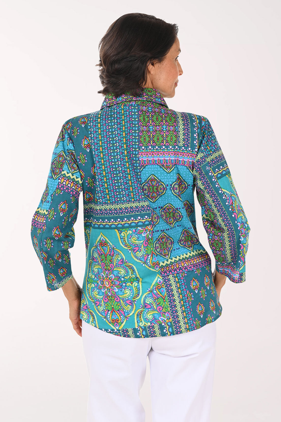 A smiling woman wearing a vibrant, multicolored patchwork print blouse with three-quarter sleeves and white pants, photographed against a solid light background.