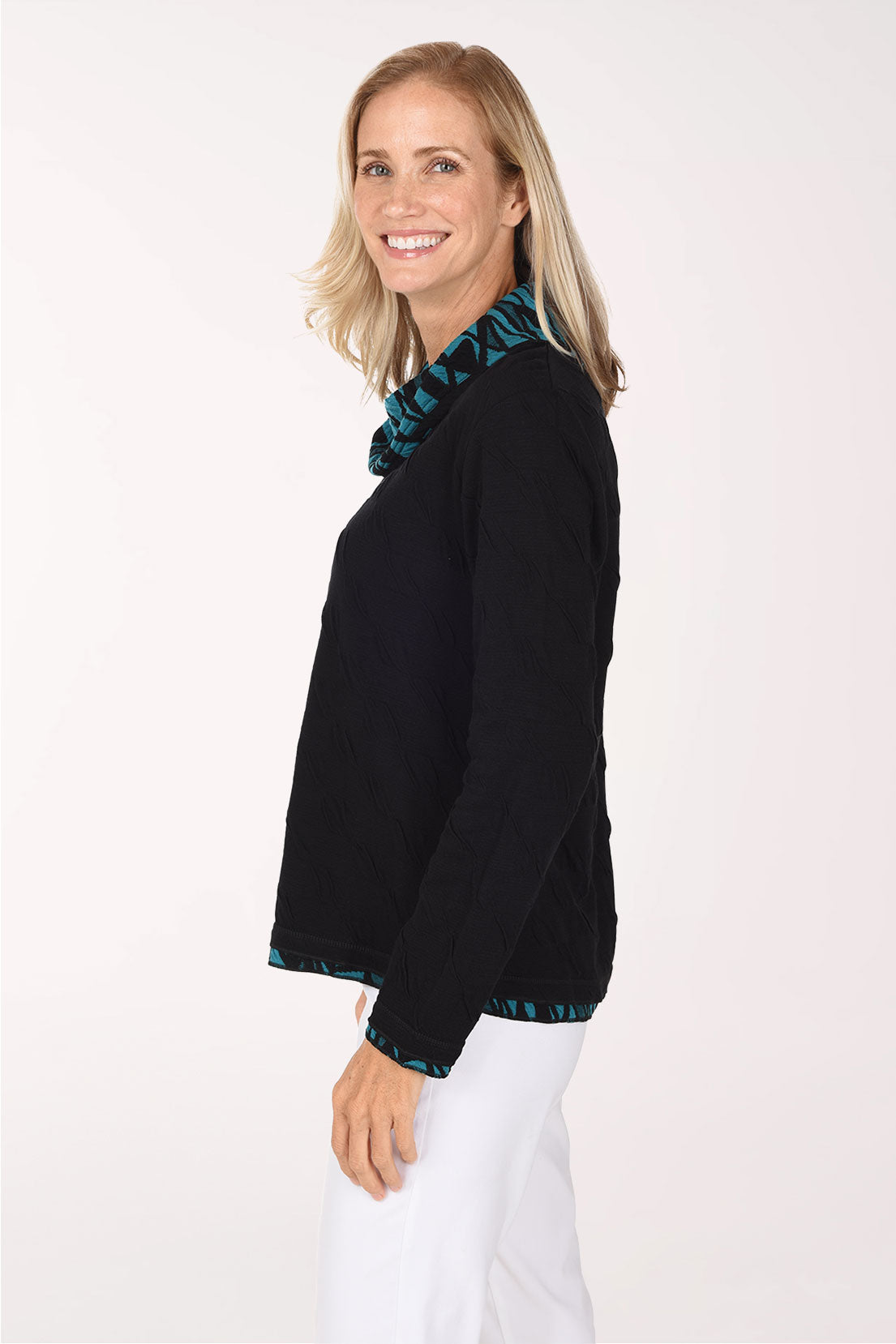 A smiling blonde woman wearing a black long-sleeve top with a teal and black patterned ruffled collar and hem, paired with white pants, against a plain white studio background.