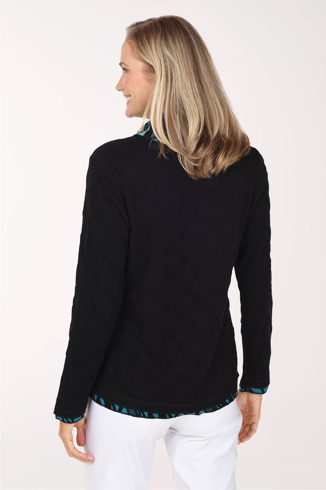 A smiling blonde woman wearing a black long-sleeve top with a teal and black patterned ruffled collar and hem, paired with white pants, against a plain white studio background.