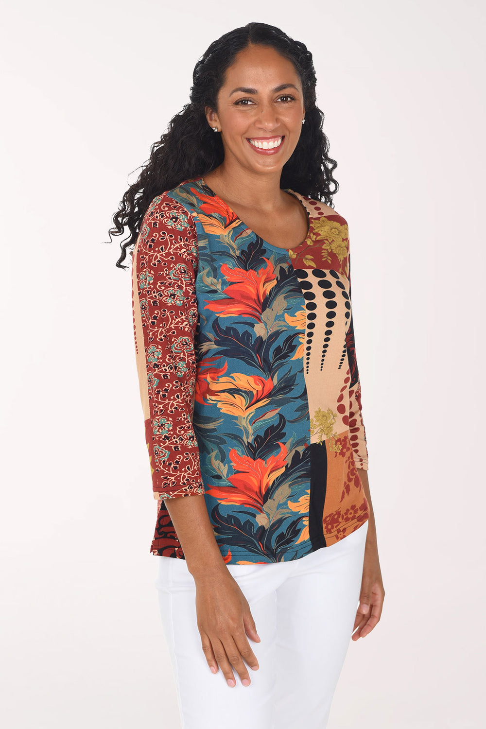 A smiling woman with long curly black hair models a three-quarter sleeve top with a multicolor abstract and floral print, paired with white pants, against a plain white studio background.
