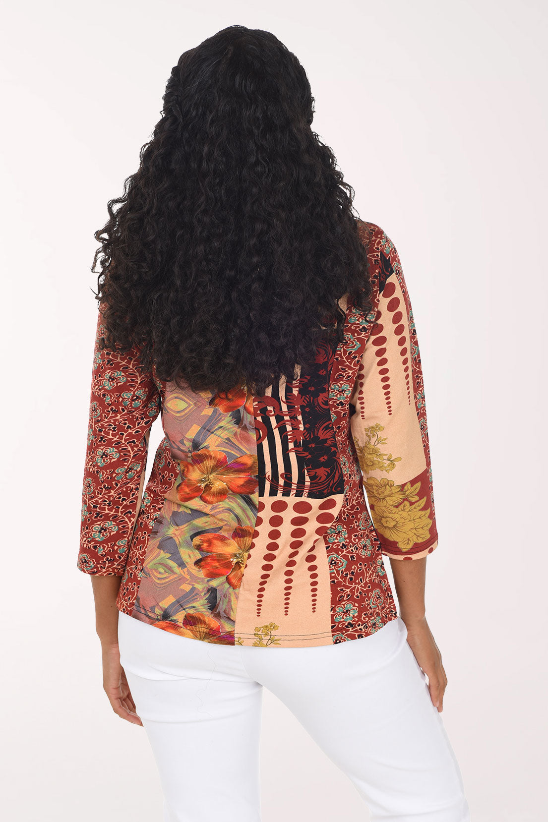 A smiling woman with long curly black hair models a three-quarter sleeve top with a multicolor abstract and floral print, paired with white pants, against a plain white studio background.
