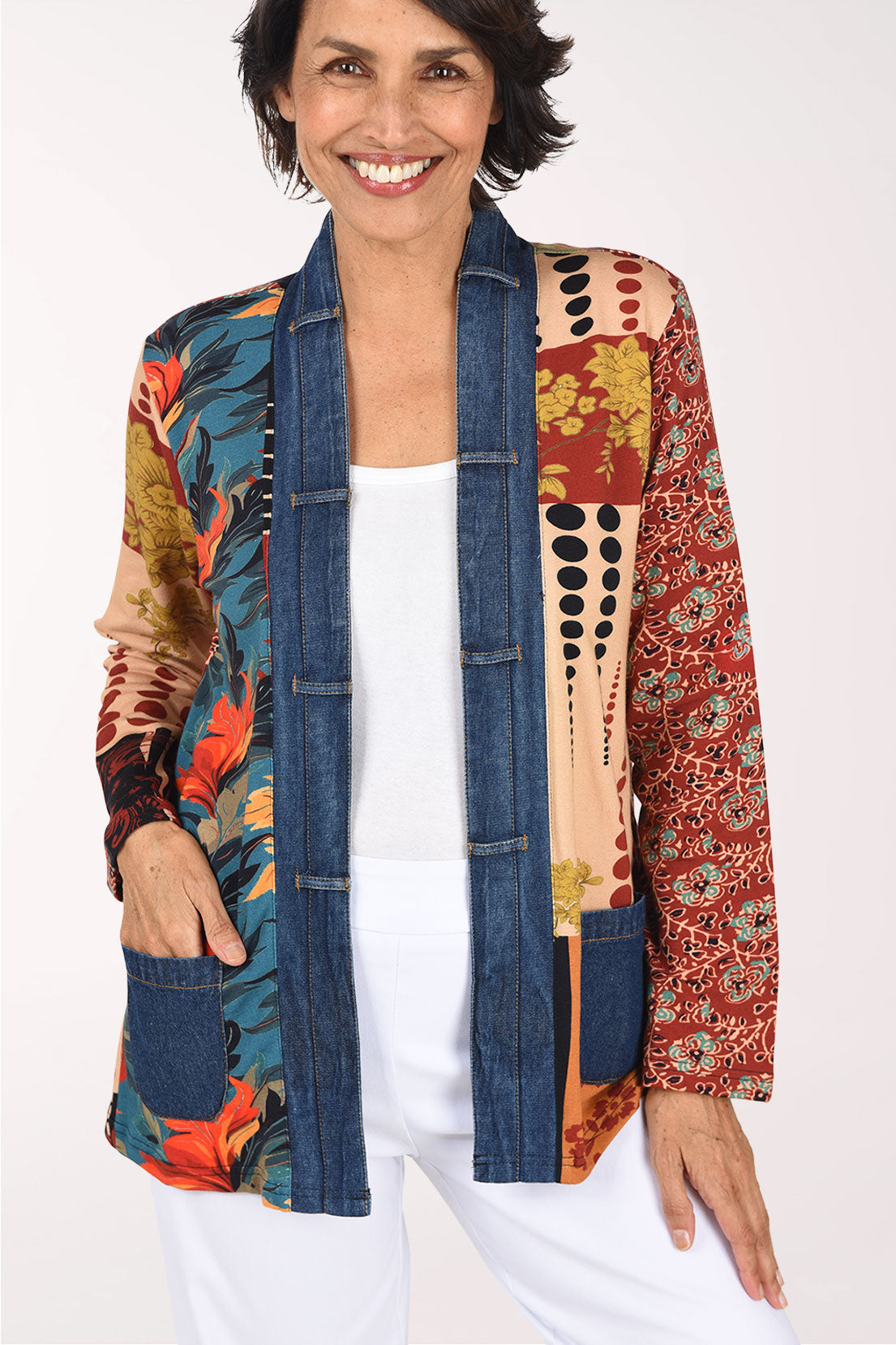 A smiling woman wearing a colorful patchwork open-front jacket with blue denim-style front panels and multicolor tropical and floral patterns on the sleeves and sides, paired with a white top and white pants, against a plain light background.