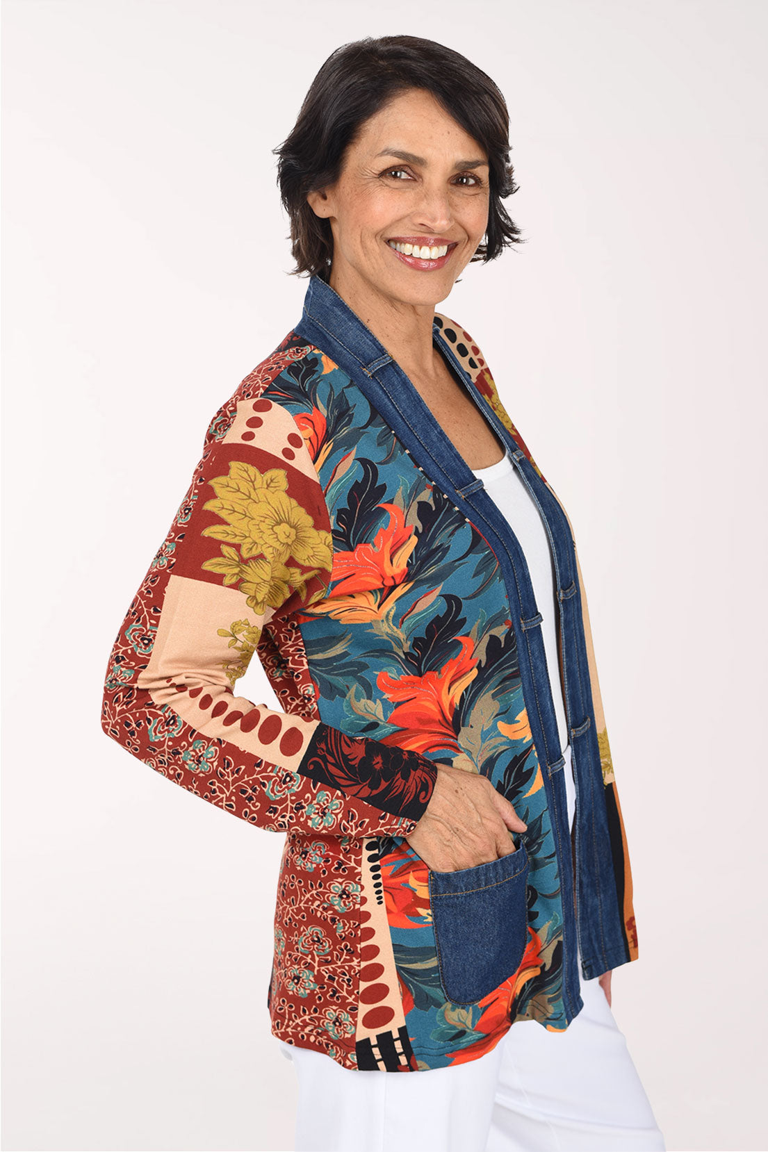 A smiling woman wearing a colorful patchwork open-front jacket with blue denim-style front panels and multicolor tropical and floral patterns on the sleeves and sides, paired with a white top and white pants, against a plain light background.