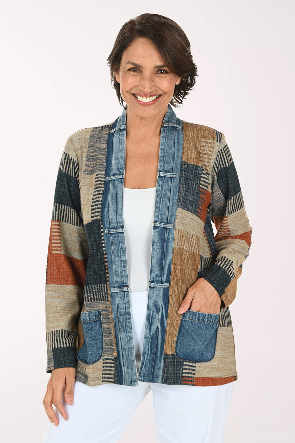 A woman modeling a patchwork cardigan with a mix of blue denim, beige, brown, and rust-orange geometric patterns, worn over a white top and white pants against a light studio background.