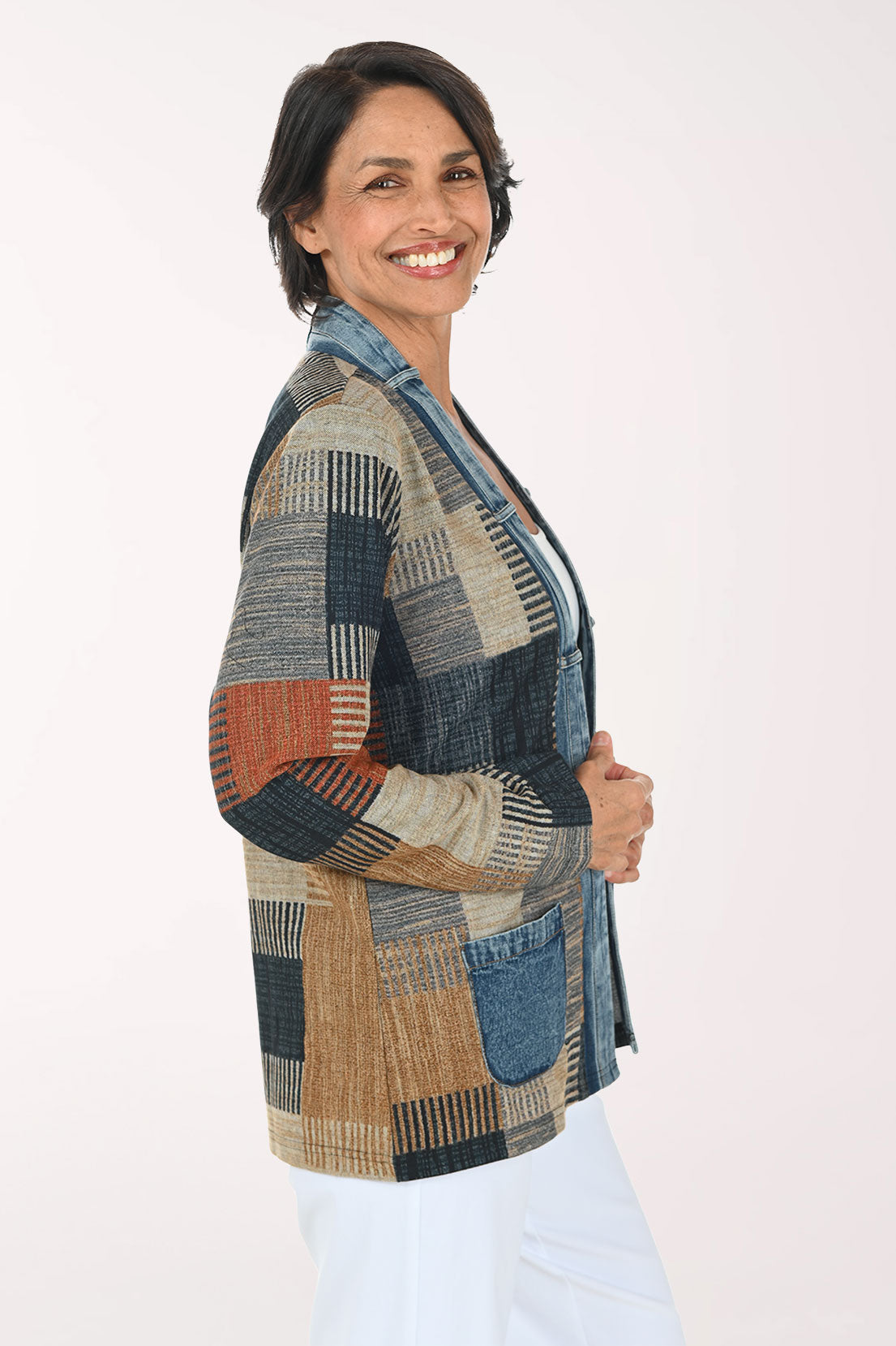 A woman modeling a patchwork cardigan with a mix of blue denim, beige, brown, and rust-orange geometric patterns, worn over a white top and white pants against a light studio background.