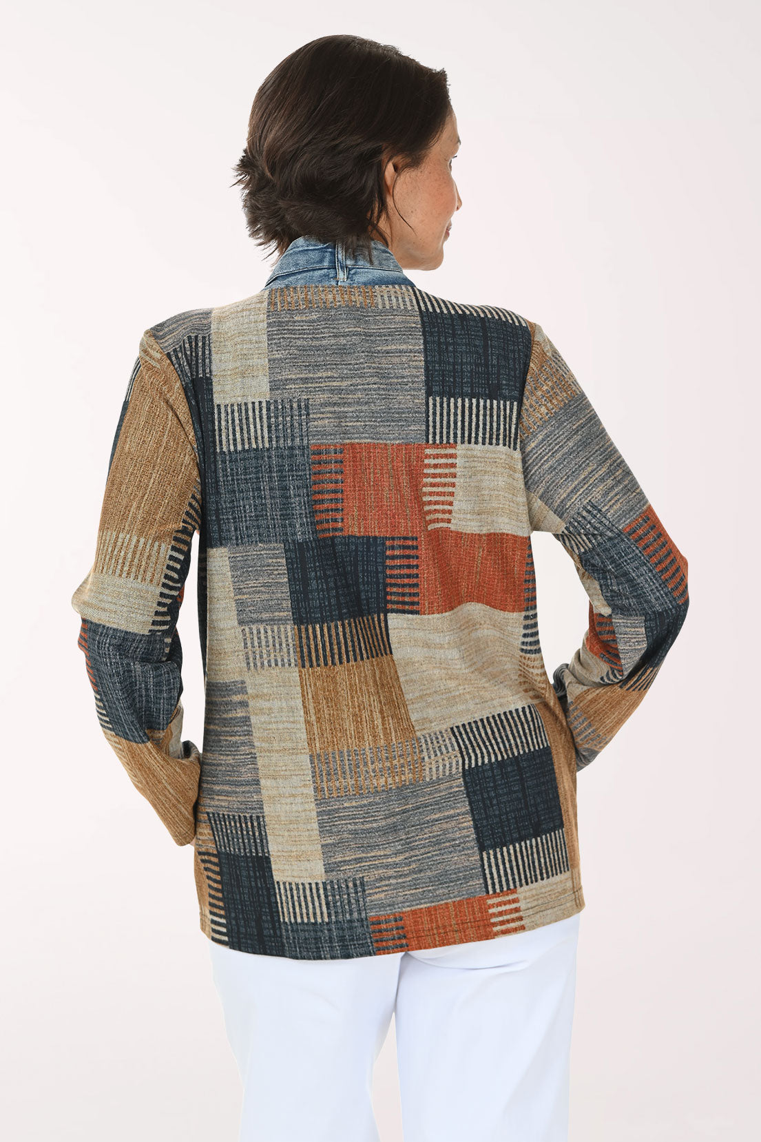 A woman modeling a patchwork cardigan with a mix of blue denim, beige, brown, and rust-orange geometric patterns, worn over a white top and white pants against a light studio background.