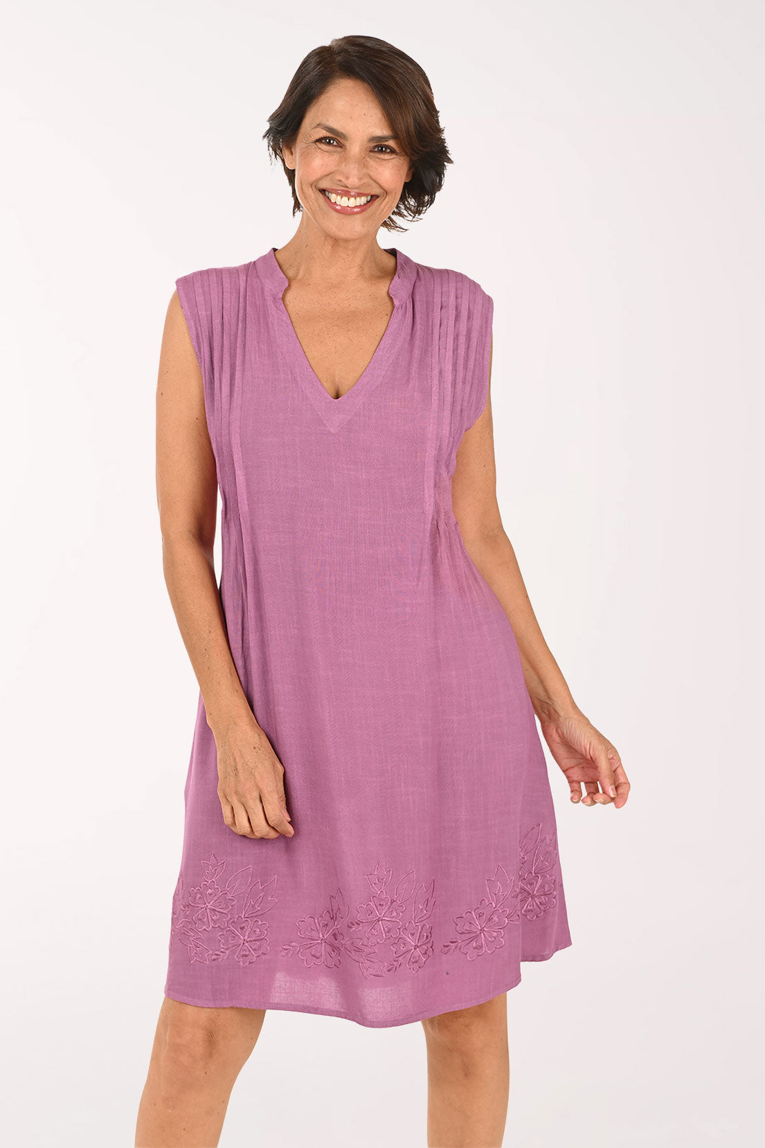 A woman modeling a sleeveless, V-neck, knee-length dress in a dusty mauve-purple color, featuring a gathered waist and decorative embroidered lace trim at the hem, set against a plain white studio background.