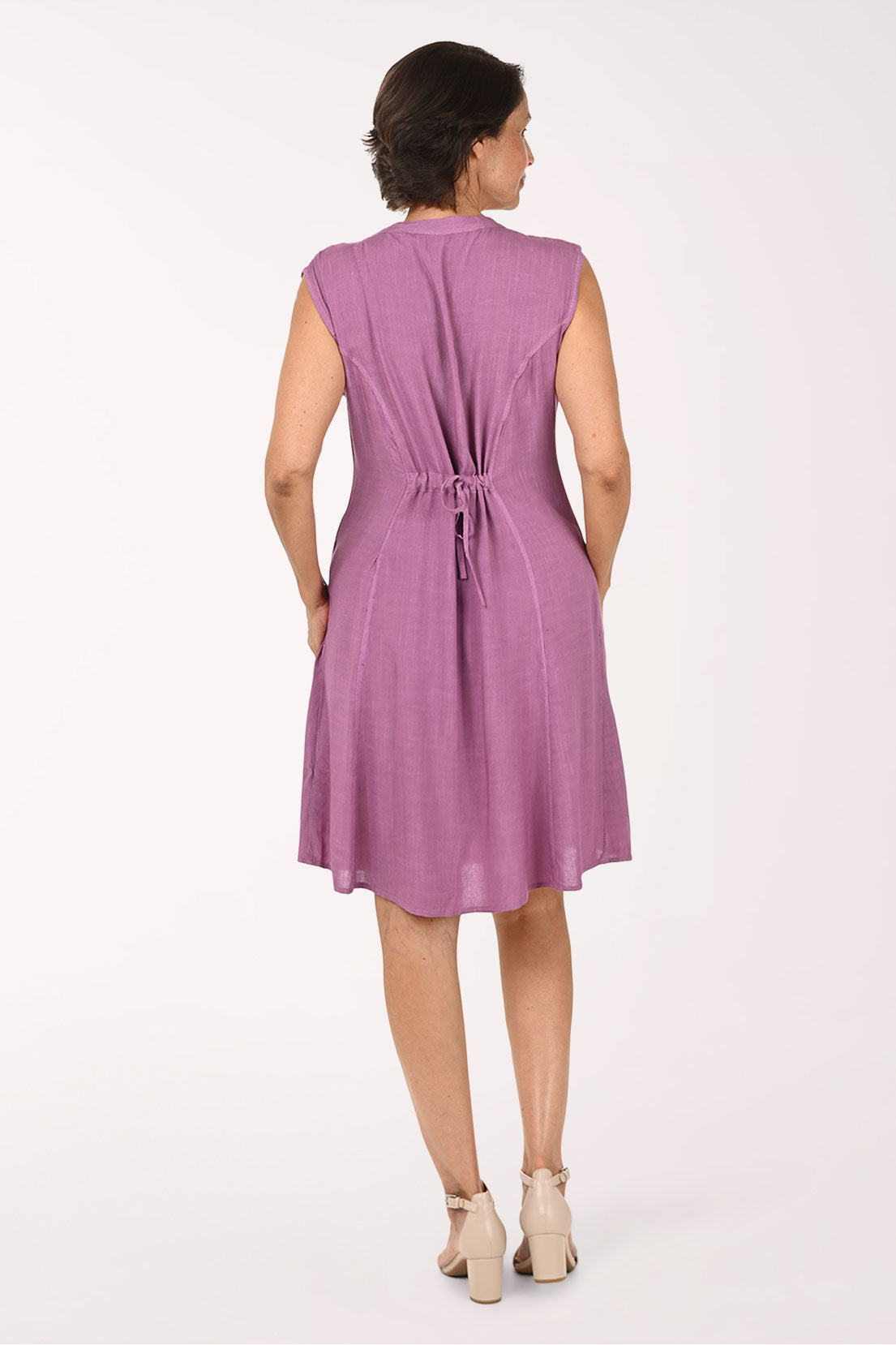 A woman modeling a sleeveless, V-neck, knee-length dress in a dusty mauve-purple color, featuring a gathered waist and decorative embroidered lace trim at the hem, set against a plain white studio background.