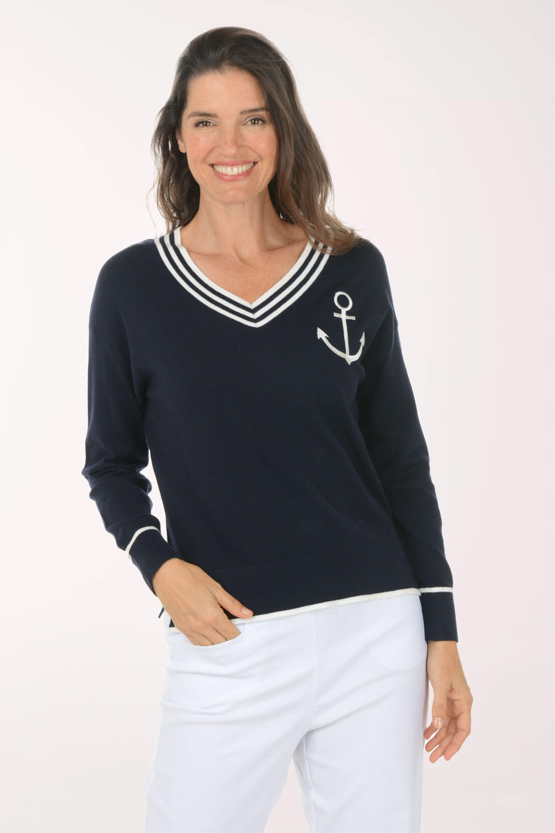 A woman modeling a navy blue long-sleeve knit sweater with a white V-neck trim, white sleeve cuffs, and a white anchor graphic on the left chest, paired with white pants against a light studio background.