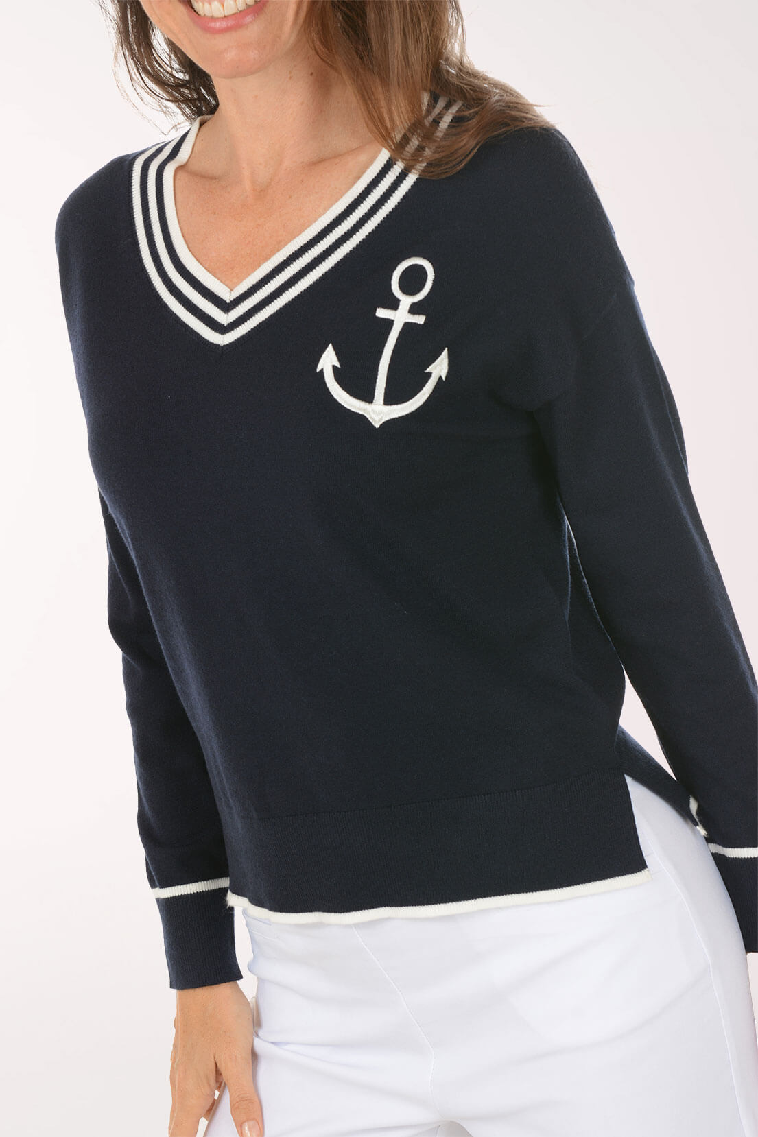 A woman wearing a navy blue long-sleeve sweater with a white V-neck trim, white stripes, and a white anchor graphic on the chest, paired with white pants.
