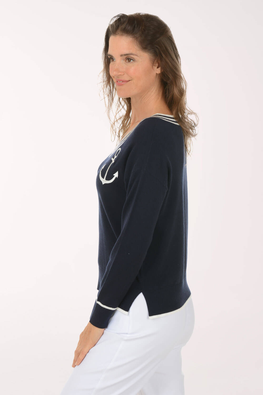 A woman shown in side profile wearing a navy long-sleeve knit top with white trim and a white anchor graphic on the chest, paired with white pants, against a plain white studio background.