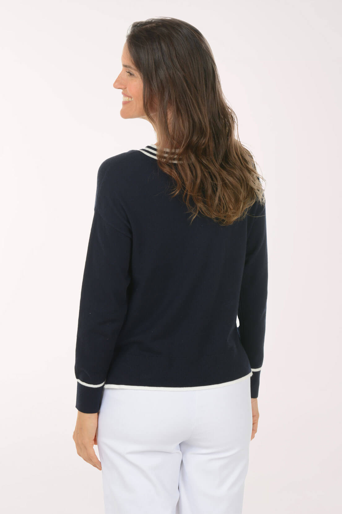 A woman shown from the back wearing a navy blue long-sleeve knit top with white stripe trim at the collar and cuffs, paired with white pants, against a plain white studio background.