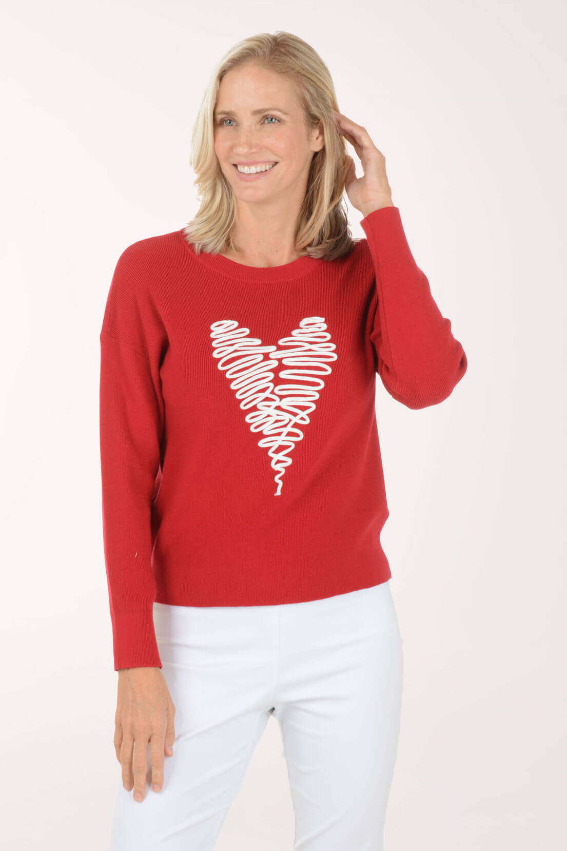 A smiling blonde woman wearing a red long-sleeve crewneck sweater with a white scribbled heart graphic on the front, paired with white pants, against a plain white studio background.