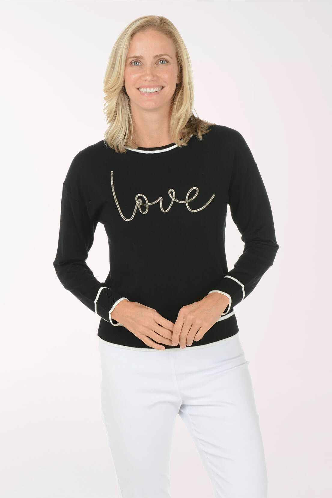 A smiling blonde woman wearing a black long-sleeve crewneck sweater with white trim and the word 'love' in cursive across the chest, paired with white pants, against a plain light background.