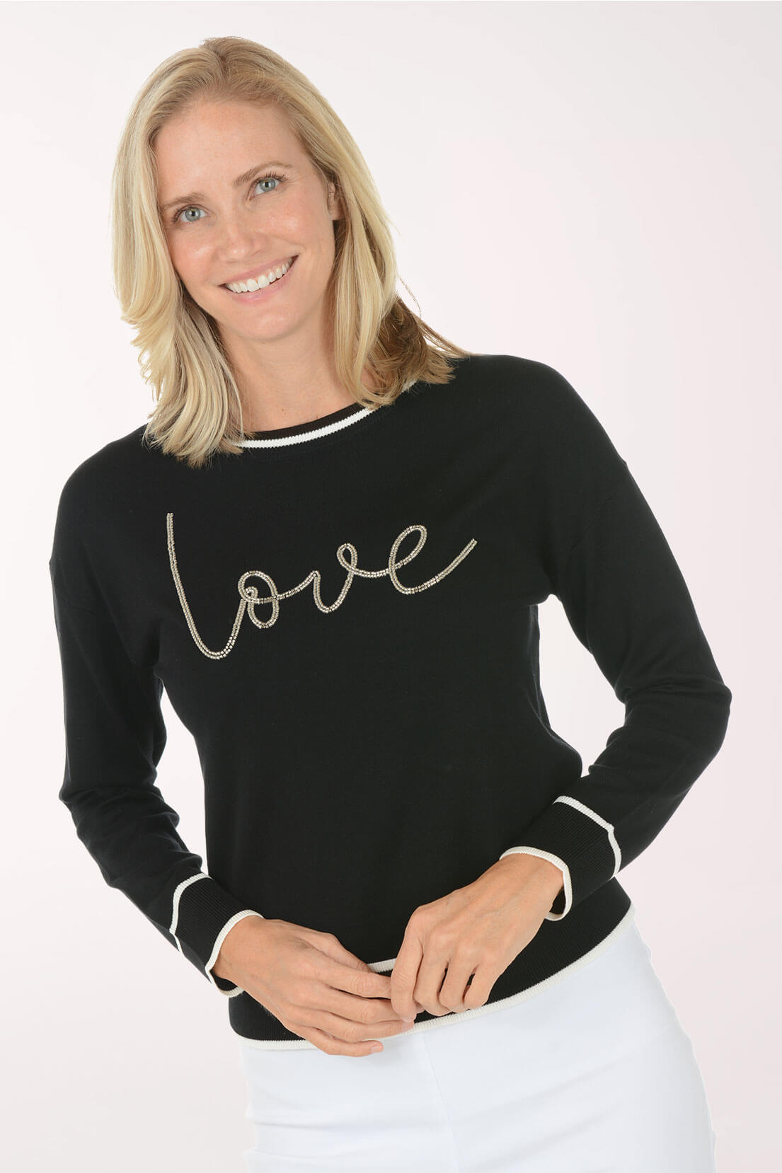 A smiling blonde woman wearing a black long-sleeve sweater with 'love' written across the chest in white script and white trim, paired with white pants, against a light studio background.