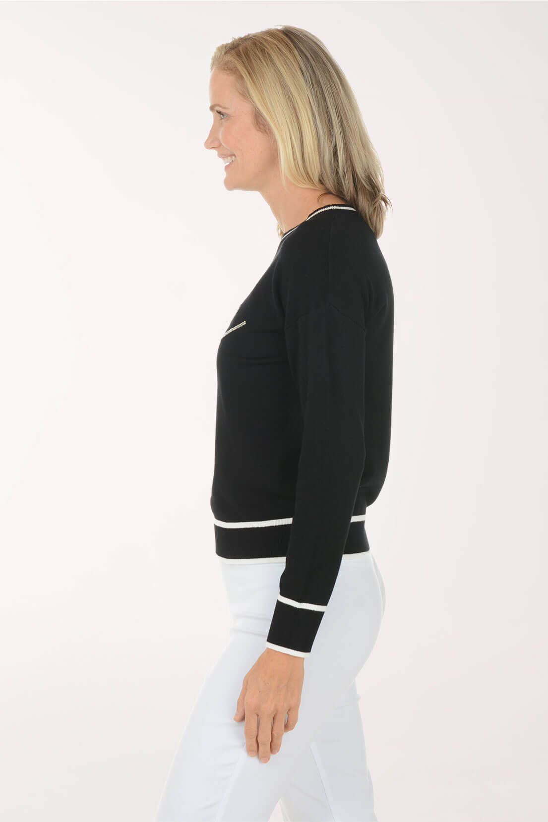 A woman shown in side profile wearing a black long-sleeve knit sweater with white contrast trim at the collar, hem, and cuffs, paired with white pants, against a plain white studio background.