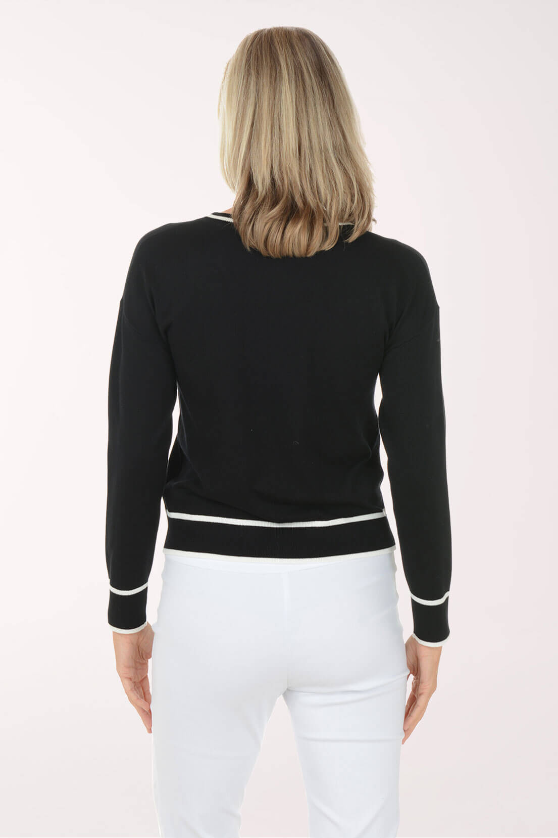 A woman viewed from the back wearing a black long-sleeve knit top with white contrast trim at the collar, cuffs, and hem, paired with white pants, against a plain light background.