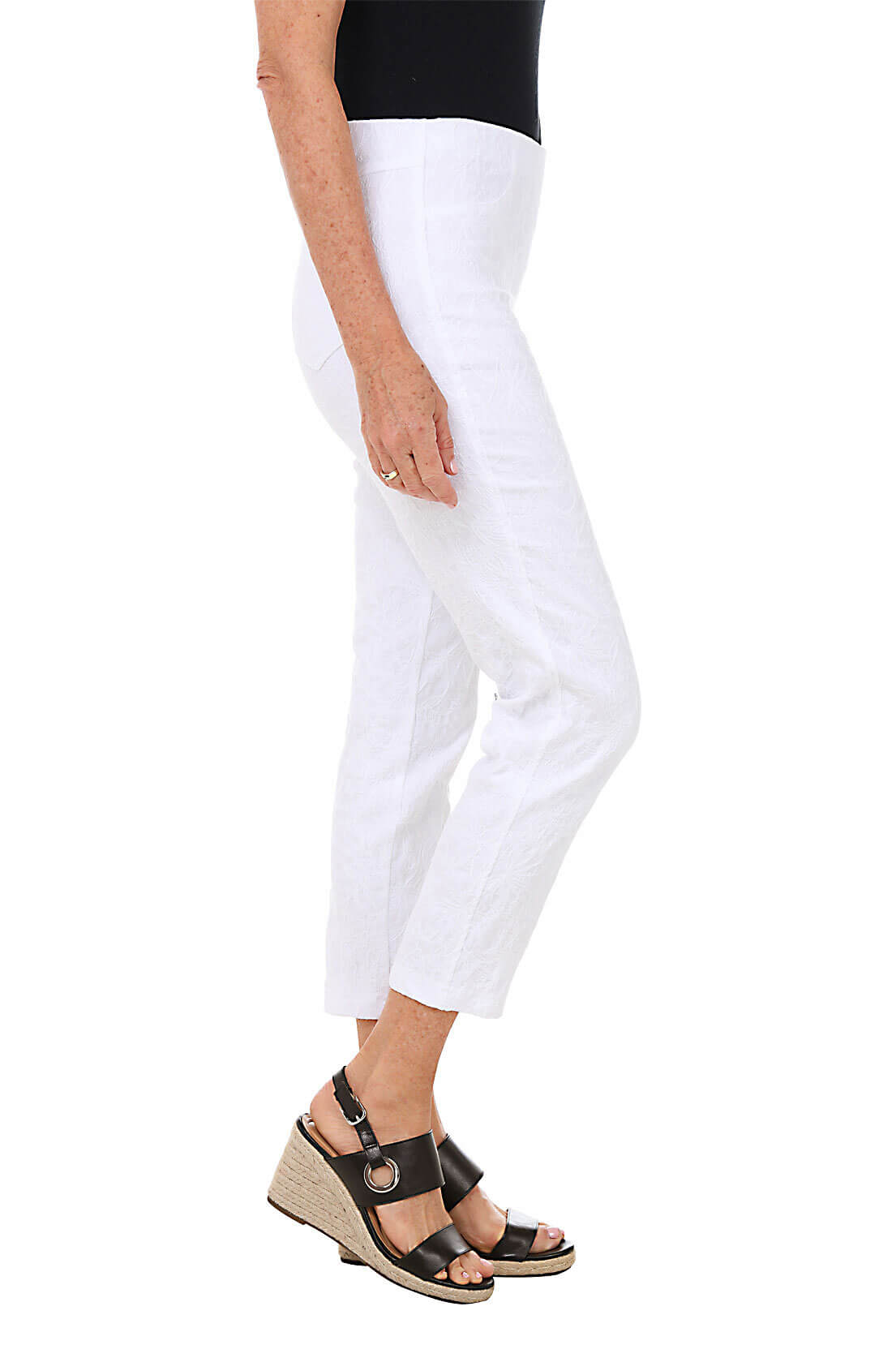 A side shot of a model wearing a pair of white floral jacquard pull-on ankle pants with faux front pockets and back patch pockets, standing against a plain background.