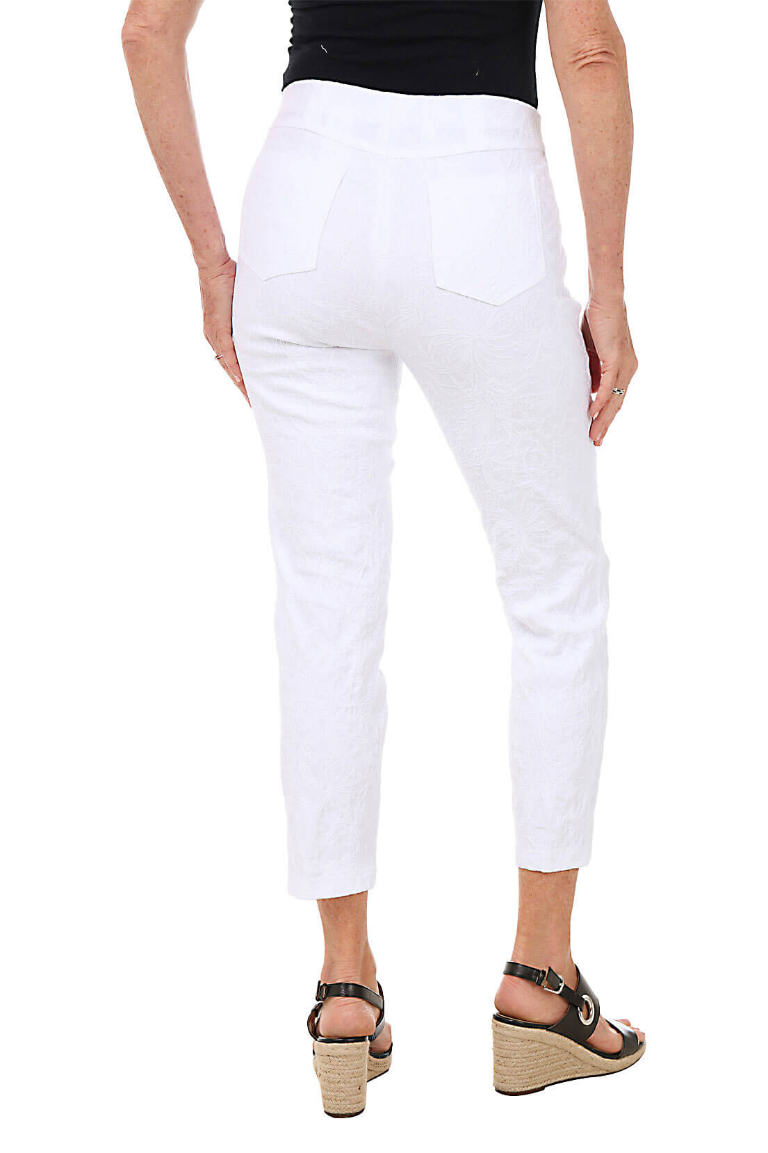 A back shot of a model wearing a pair of white floral jacquard pull-on ankle pants with faux front pockets and back patch pockets, standing against a plain background.