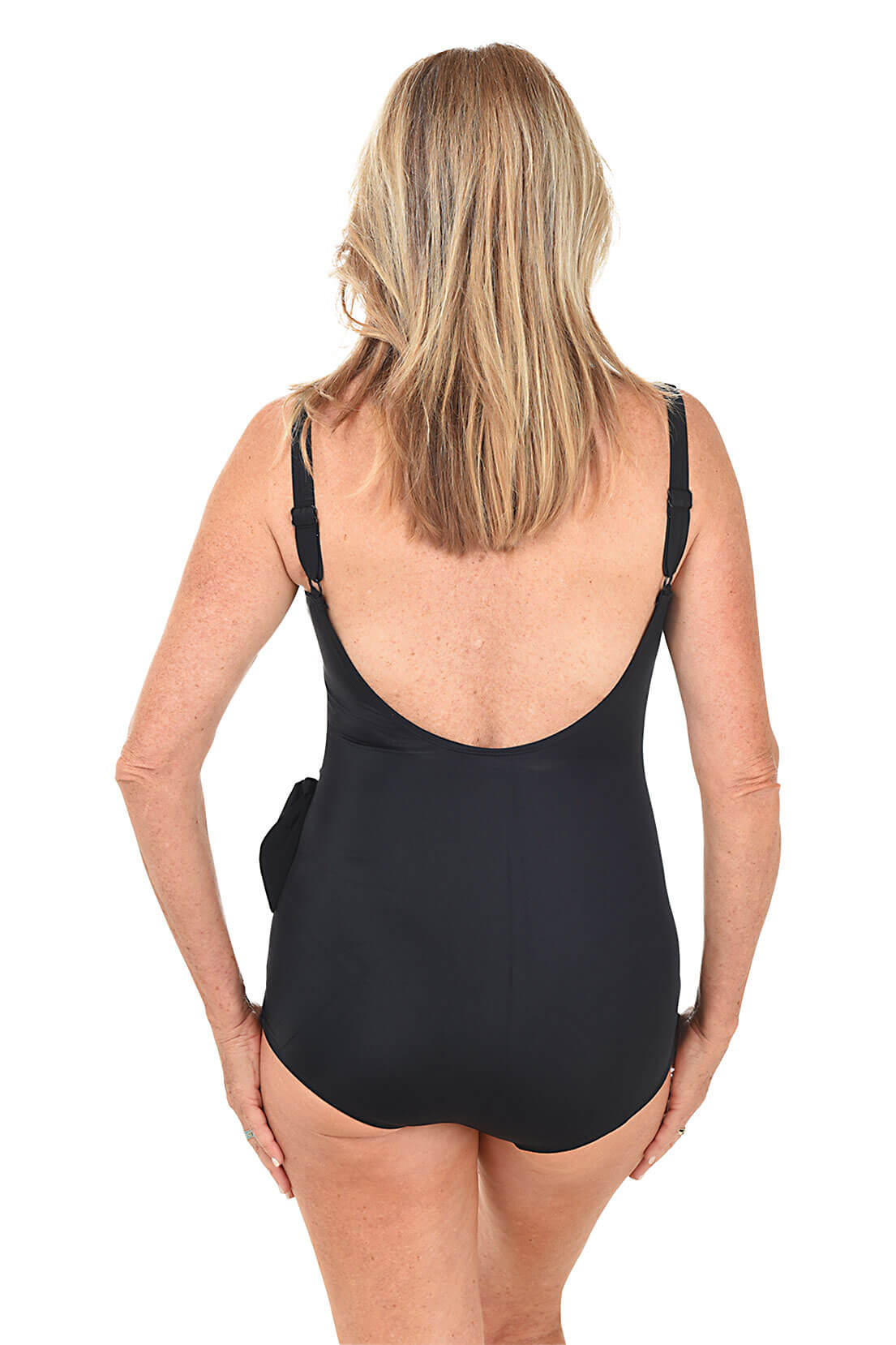 A back shot of a model wearing a solid black v-neck sarong swimsuit with adjustable straps and a decorative tie on the left side.