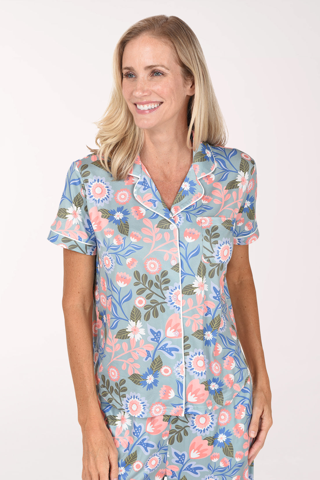 A smiling blonde woman wearing a short-sleeve pajama set with a light blue background and a colorful floral print, standing against a plain white studio background.