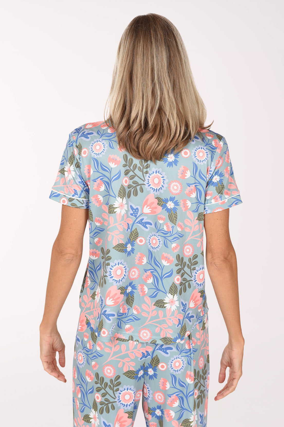 A smiling blonde woman wearing a short-sleeve pajama set with a light blue background and a colorful floral print, standing against a plain white studio background.