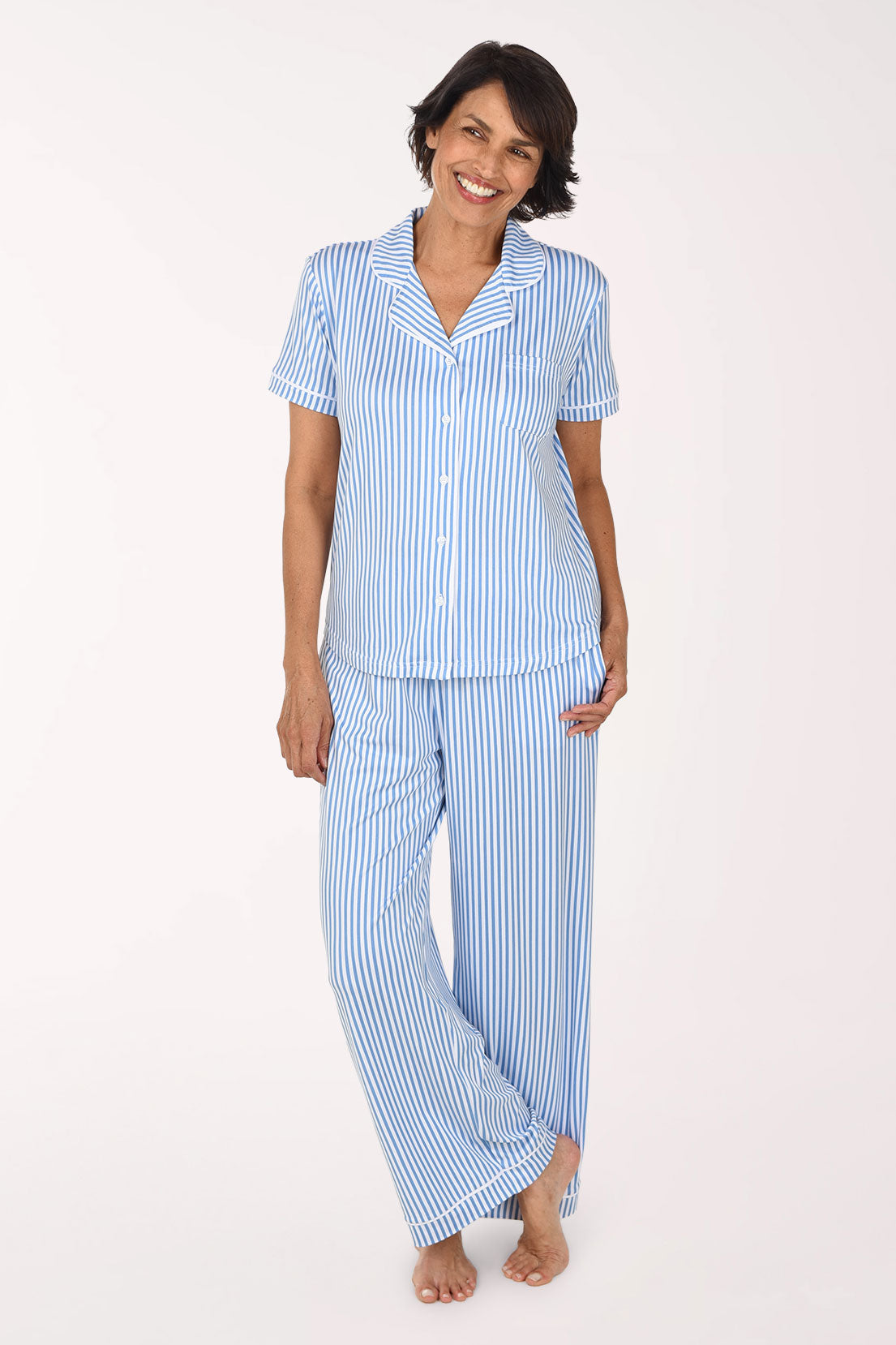 A woman modeling a two-piece pajama set with light blue and white vertical stripes, featuring a short-sleeve button-front top and matching long pants, against a plain white studio background.