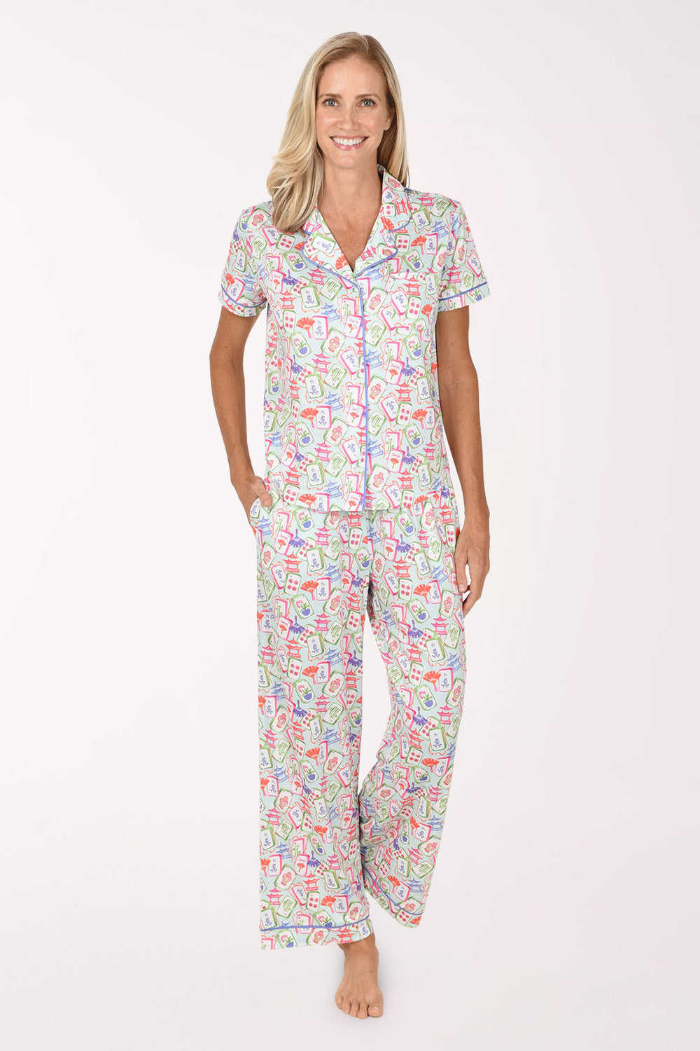 A woman modeling a matching short-sleeve button-up pajama shirt and wide-leg pajama pants set with a colorful tropical print on a white background.