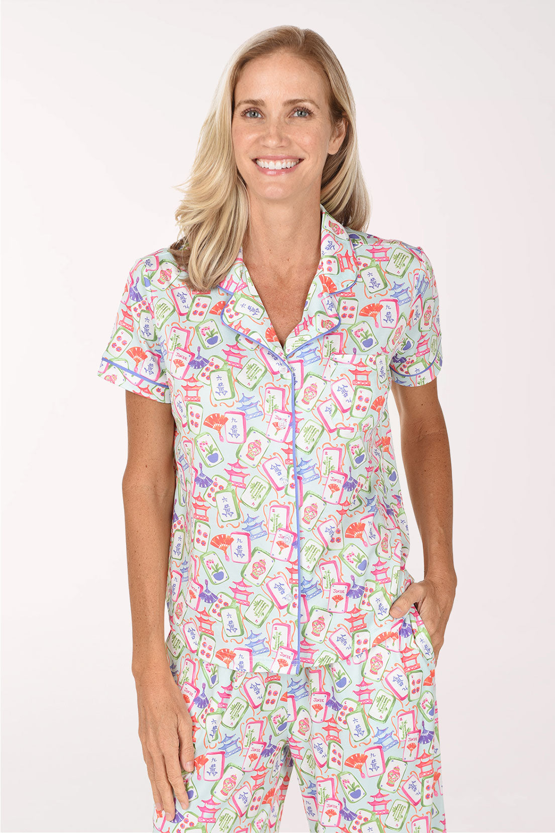 A smiling blonde woman wearing a short-sleeve button-front pajama top and matching pants with a colorful cartoon-style print on a white base, photographed against a plain light background.